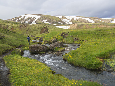 云纹摄影照片_孤独的徒步旅行者在冰岛自然保护区 Fjallabaki 的 f210 路附近的地热区 Strutslaug 的野溪流、绿草苔藓草甸和积雪覆盖的流纹岩山脉中行走
