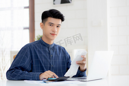 年轻的亚洲男子用家里的办公桌上的计算器计算财务家庭，男性检查账单和成功节省家庭开支，债务清偿，税务和会计，商业概念。