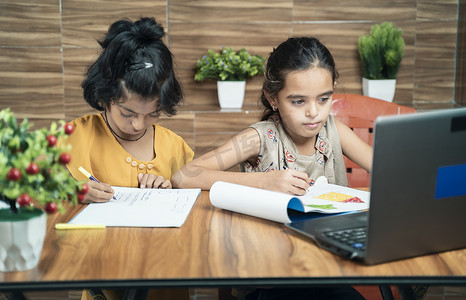 两个孩子在家里的在线或虚拟课堂上看着笔记本电脑忙着写作 — 冠状病毒或 covid-19 大流行期间电子学习或远程学习或儿童生活方式的概念