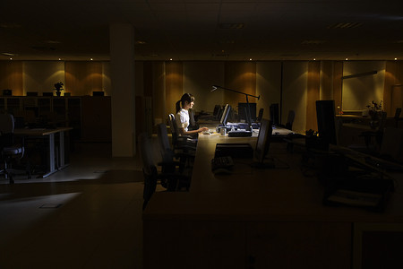 一位在黑暗办公室用电脑工作的年轻女性的侧视图
