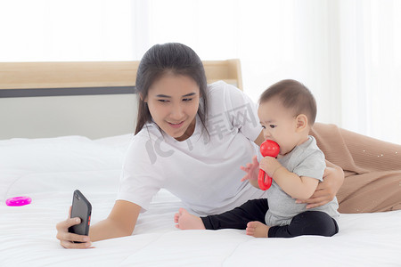 年轻的亚洲母亲和小女婴或新生儿在卧室床上用智能手机自拍，幸福的妈妈和女儿在家里使用电话视频通话，两个人，家庭和沟通概念。