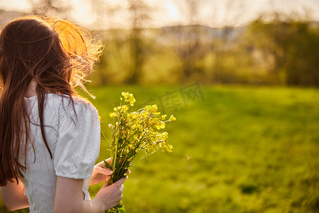 日落时分，一位身穿白色裙子、留着长红头发的女士站在绿色田野中，背对着镜头