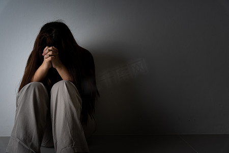 哭泣摄影照片_悲伤的女人抱着她的膝盖，独自坐在黑暗的房间里哭泣。