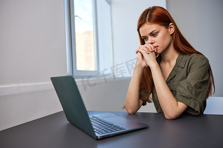 双手交叉摄影照片_一位表情严肃、工作疲惫的女性坐在笔记本电脑前，双手交叉放在脸旁