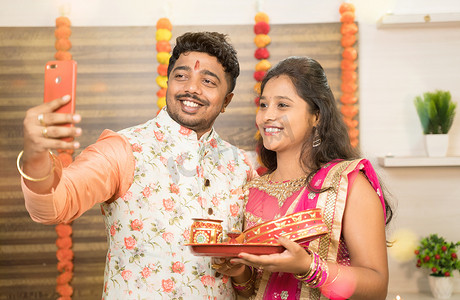 节后错峰游摄影照片_身着传统服饰的印度夫妇在家里庆祝 Karva Chauth 节后自拍 — 在节日庆祝活动中使用移动技术的概念。