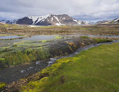 在冰岛自然保护区 Fjallabaki 的 f210 路附近的地热区，夏季景观与五颜六色的绿色和橙色苔藓草甸与热水池 Strutslaug 和春天与白雪覆盖的流纹岩山脉