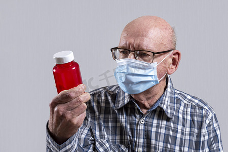 戴眼镜的老头摄影照片_秃头老头戴着眼镜，戴着医用面具，手里拿着一罐红色的药丸。