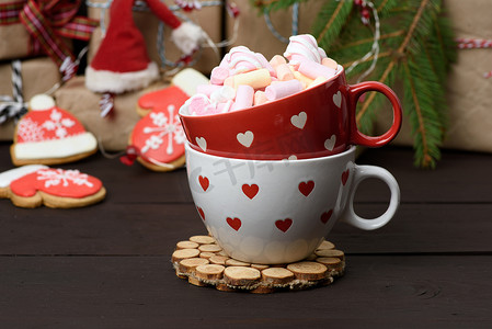 圣诞花环摄影照片_带可可和棉花糖的红色陶瓷杯，放在礼盒和圣诞花环后面