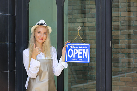 自信的年轻女性商店的肖像，小企业咖啡店的老板站在店前，上面有开放的标志。