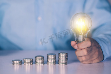 商人的手拿着灯泡，同时在办公桌上投币，为成功储蓄和融资，关于存款和收入和投资的知识，解决方案和灵感，商业理念。