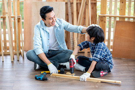亚洲父亲在家庭木工工作场所钻木头时，心情愉快地摸着儿子的头。