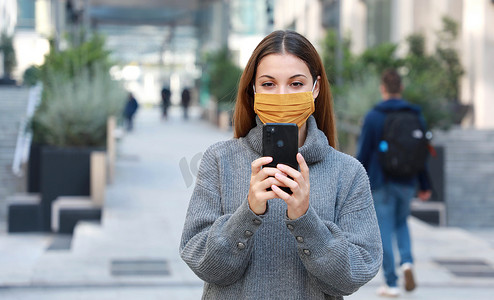 戴着织物面具的年轻女子在步行街上行走时用手机发短信。