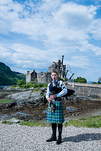 城堡城堡摄影照片_蓝天背景的苏格兰高地著名艾琳多南城堡前的风笛演奏者