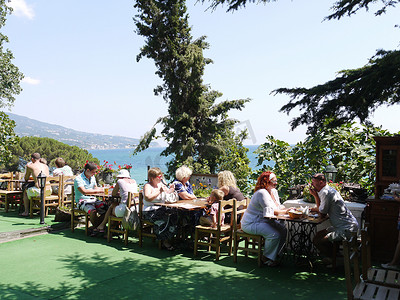来到海边咖啡馆的游客坐在被绿树环绕的桌子旁，可以俯瞰大海、海边的青山和万里无云的蓝天。