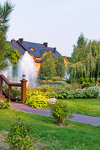 湖中的喷泉位于一座大型木屋前，周围环绕着绿色公园、草坪和树木，背景是淡蓝色的天空。