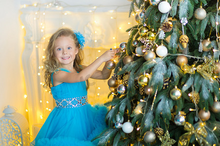 年轻漂亮的女孩穿着蓝白色优雅的晚礼服坐在圣诞树附近的地板上，并在新年前夜礼物。