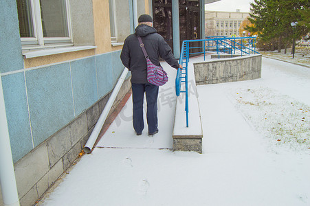 一位白发苍苍的老人在积雪覆盖的残疾人坡道上艰难攀爬，在雪中留下痕迹