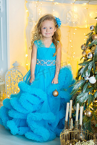 蝴蝶节摄影照片_年轻漂亮的女孩穿着蓝白色优雅的晚礼服坐在圣诞树附近的地板上，并在新年前夜礼物。