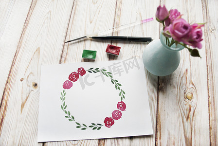 玫瑰花环手绘摄影照片_艺术家工作区配有水彩、画笔、玫瑰花束、木制背景上手绘花环框。