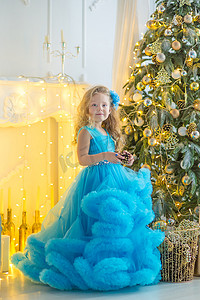 蝴蝶节摄影照片_年轻漂亮的女孩穿着蓝白色优雅的晚礼服坐在圣诞树附近的地板上，并在新年前夜礼物。