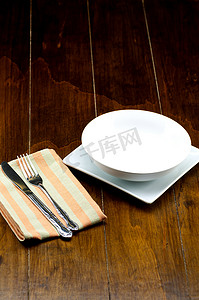 木质方形盘子摄影照片_方形盘子上的空碗，餐巾上有叉子和刀子，在木质背景上。