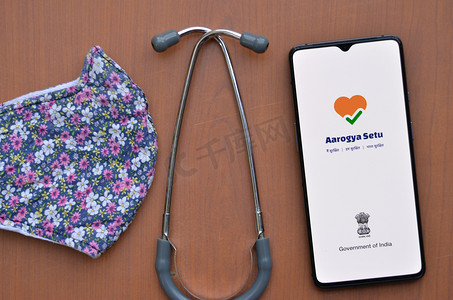 政务摄影照片_印度新德里，2020 年。平躺着手机，展示 Aarogya Setu 应用程序，带有听诊器和设计师花卉面罩木制背景。