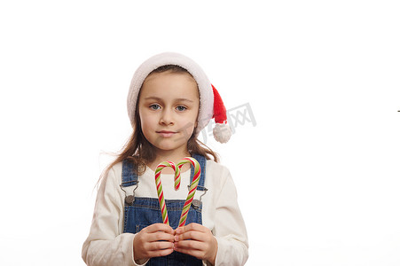 齿形边框摄影照片_可爱的蓝眼睛小女孩戴着圣诞老人的帽子，拿着心形的拐杖糖。