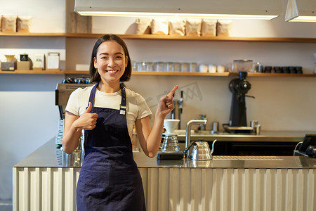 快乐的年轻亚洲咖啡师，穿着围裙的女职员，竖起大拇指，指着复印空间，展示咖啡馆广告