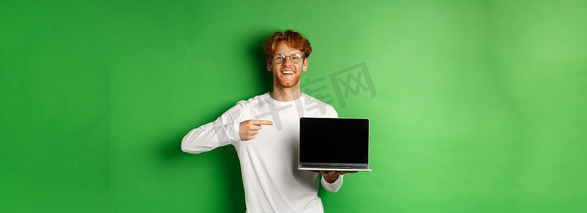 戴着眼镜和白色长袖 T 恤的快乐红发男子，用手指指着空白的笔记本电脑屏幕，微笑着，站在绿色背景上