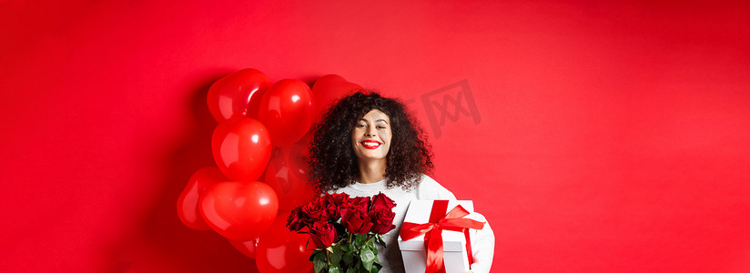 气球盒子摄影照片_微笑的幸福女人拿着盒子，里面装着男朋友的礼物和红玫瑰，庆祝情人节，站在浪漫的心形气球旁边，站在工作室背景上