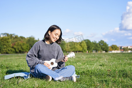 唱歌的亚洲女孩在草地上弹尤克里里琴，坐在公园的毯子上，在阳光明媚的日子在户外放松