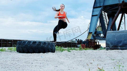 一位身穿红色上衣和黑色紧身裤的年轻运动女性使用大型重型拖拉机轮进行不同的力量练习，训练她的肌肉。