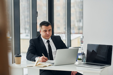 穿西装打领带的男人坐在桌子旁，拿着笔记本电脑，在办公室工作