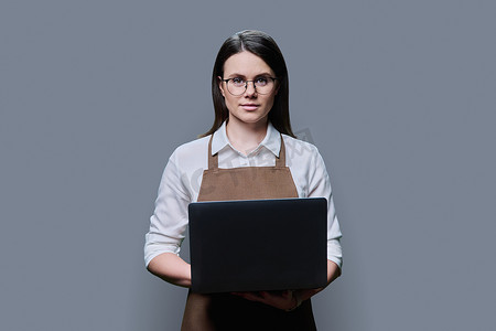 灰色工作室背景中穿着围裙的年轻女子使用笔记本电脑