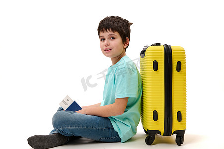 儿童白底摄影照片_帅气的多民族少年乘客，拿着登机牌，靠在黄色手提箱上，微笑着看着镜头