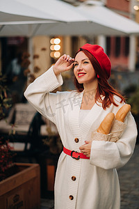 一位戴着红色贝雷帽、身穿白色羊毛衫、手里拿着法棍面包的漂亮女人在秋天的城市里漫步的肖像