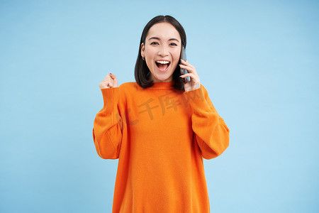 快乐的亚洲女孩通过电话收到好消息，拿着手机，握拳，庆祝胜利，成就，蓝色工作室背景