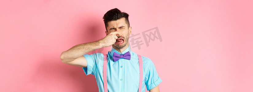 照片中，一名男子因恶心的气味而哭泣，闭上鼻子以免发出难闻的臭味，站在粉色背景的领结和吊带上