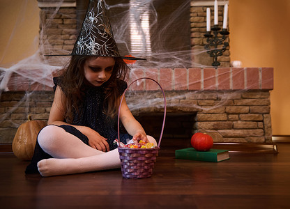 巫帽摄影照片_小女巫戴着巫师帽，坐在蜘蛛网覆盖的壁炉旁。