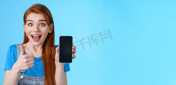 手机批准摄影照片_惊讶着迷的可爱红发女人推荐好的智能手机，很棒的新应用程序，手机游戏，显示手机显示屏，看起来兴奋的相机微笑批准，就像很酷的设备，蓝色背景