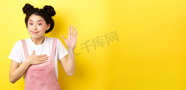 可爱的亚洲女孩化妆的形象是诚实的，举手并放在心上，说真话，宣誓和微笑，站在黄色背景上
