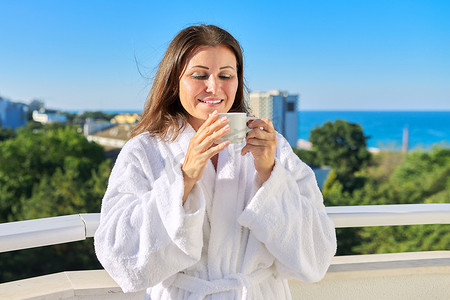海滨度假小镇酒店阳台上穿着白色浴袍的快乐中年女性