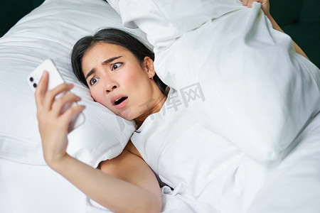 亚洲女性在床上醒来，震惊地看着手机，意识到自己睡过头了