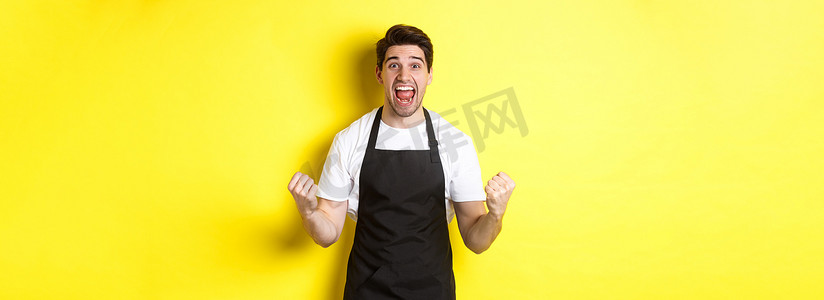 兴奋的咖啡店老板穿着黑色围裙庆祝，握拳，欢呼，实现目标，站在黄色背景下