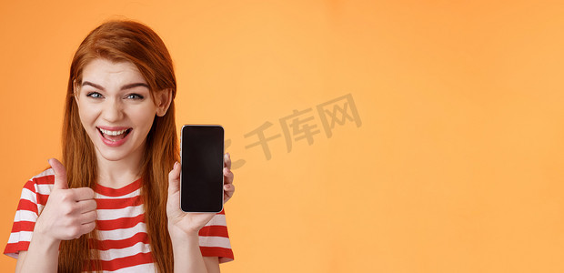 开朗自信的红发女性推荐智能手机应用程序，显示手机显示屏，竖起大拇指像批准标志，广泛微笑，建议良好的在线购买，移动广告