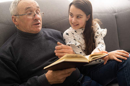 照片中，年迈的老爷爷小漂亮的孙女坐在沙发上拥抱居家隔离安全，在室内阅读有趣的书籍童话现代室内客厅。