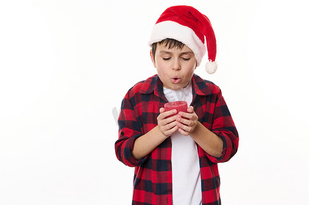 美好愿望摄影照片_穿着红色格子衬衫和圣诞帽的迷人青春期前男孩，在吹灭蜡烛的同时许下了圣诞节的美好愿望