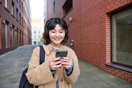 年轻韩国女性背着背包和耳机在城市漫步、听音乐、看智能手机、在街上使用手机应用程序的肖像