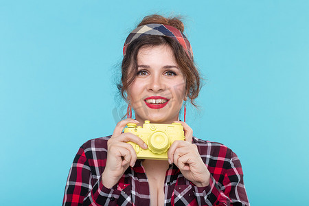 胶片格子摄影照片_一位身穿格子衬衫、手持黄色胶片老式相机、蓝色背景的漂亮年轻积极女性的画像。