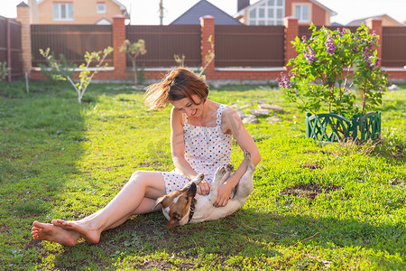 在阳光明媚的夏日，穿着裙子、笑得积极的年轻女子和她心爱的不安分的狗坐在乡间别墅的院子里玩耍。